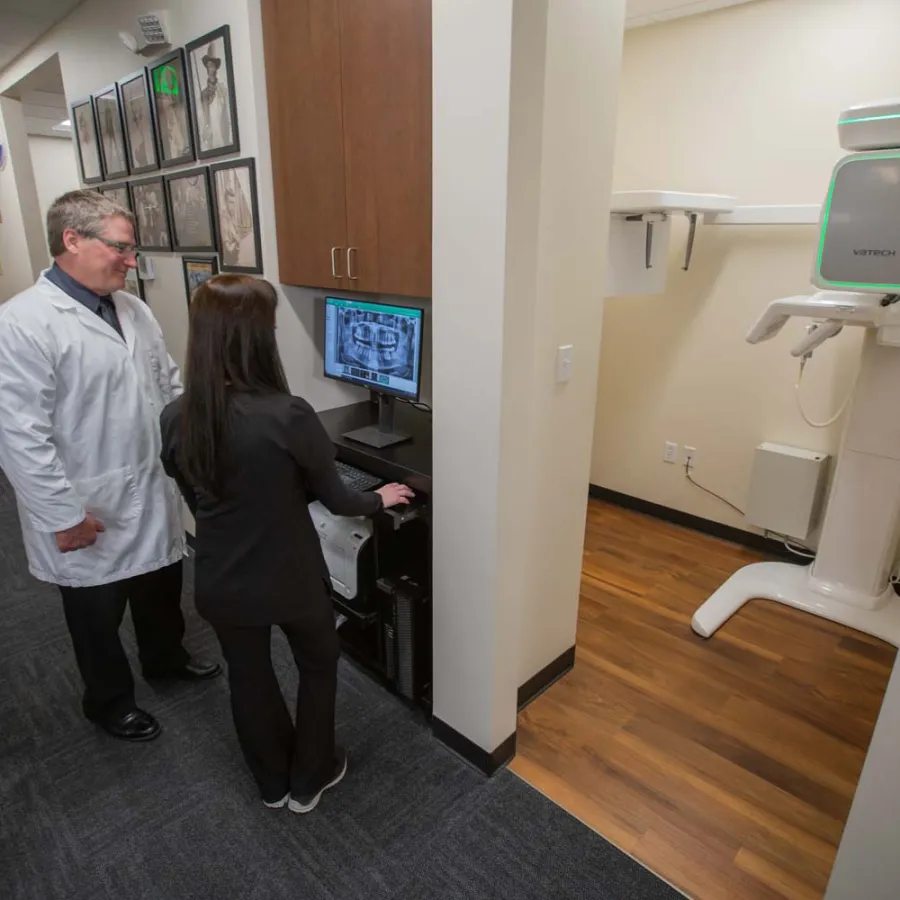 Dentist and assistant reviewing dental X-ray images on computer in modern clinic hallway near imaging equipment.