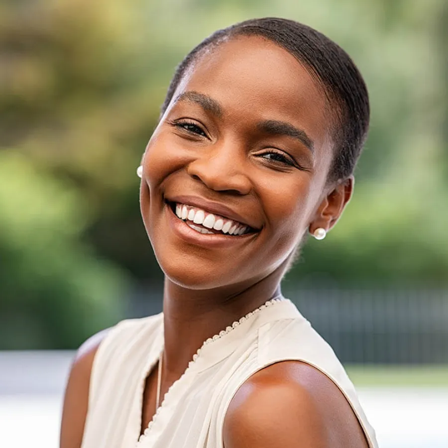 Smiling young woman with smooth skin and pearl earrings in a bright outdoor setting.