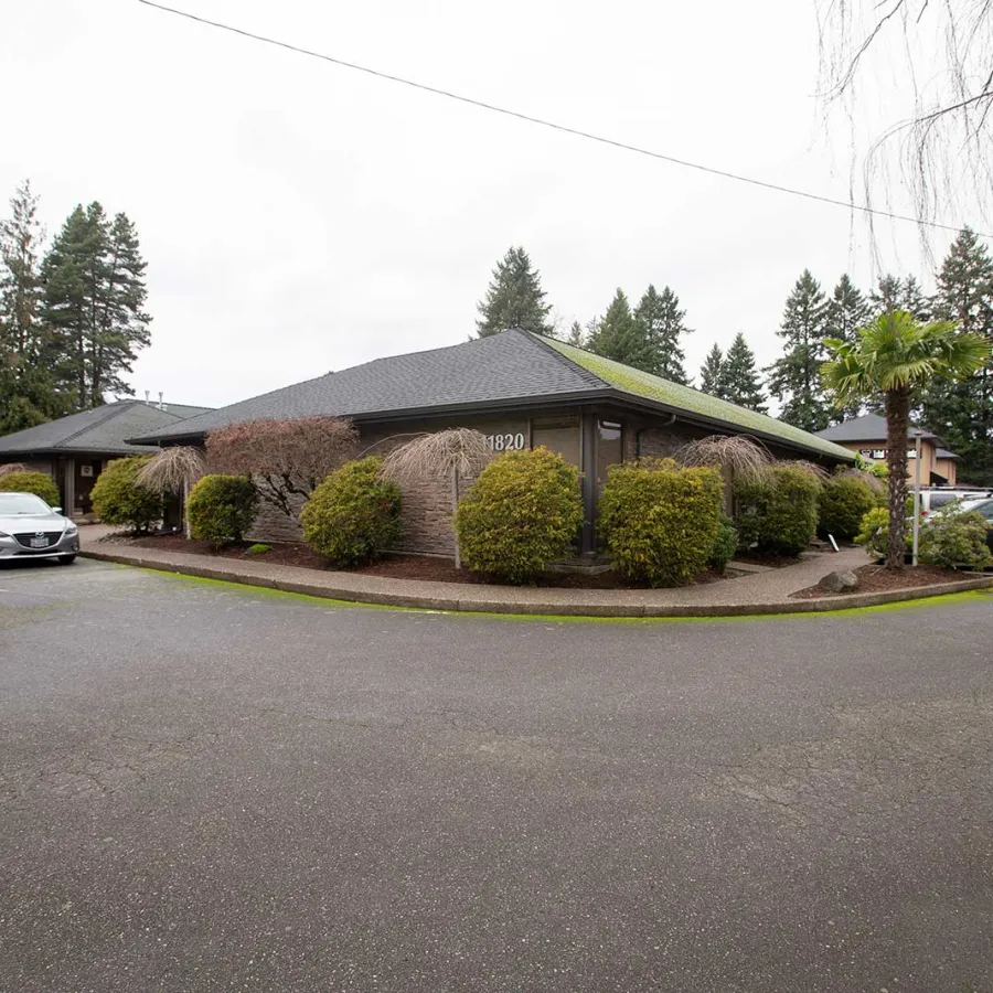 Single-story commercial building with shrubs, parked cars, and tall pine trees under an overcast sky.