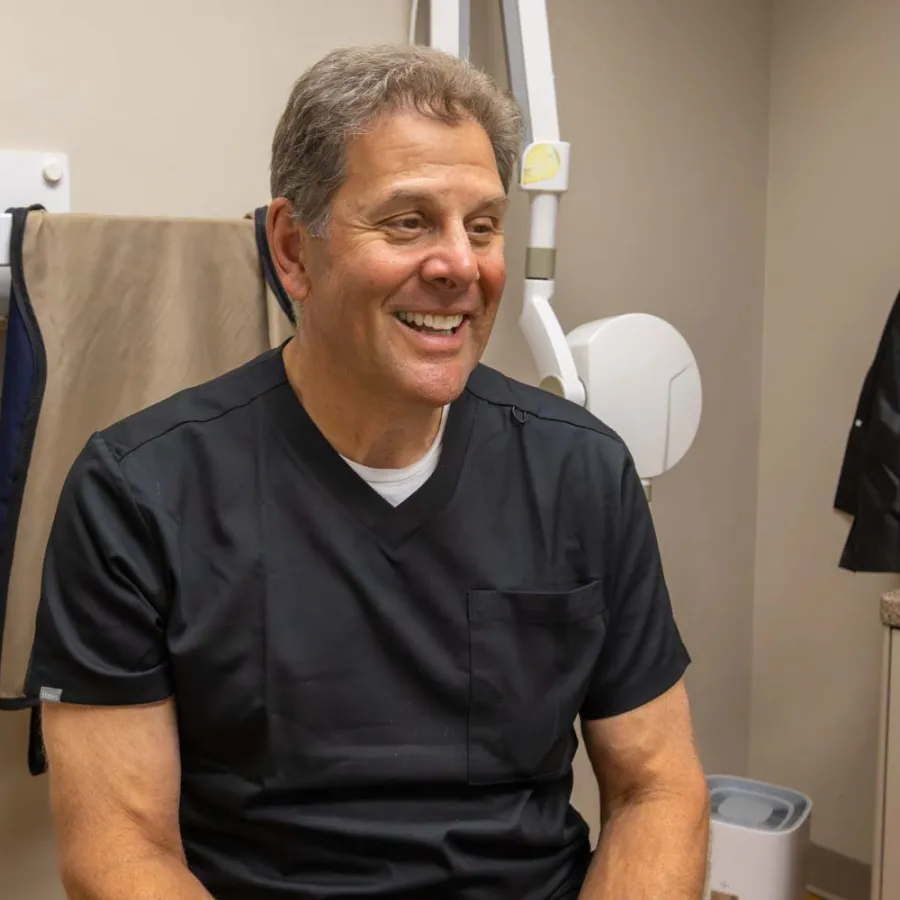 Smiling male doctor in black scrubs sitting in a medical office with equipment and coats in the background