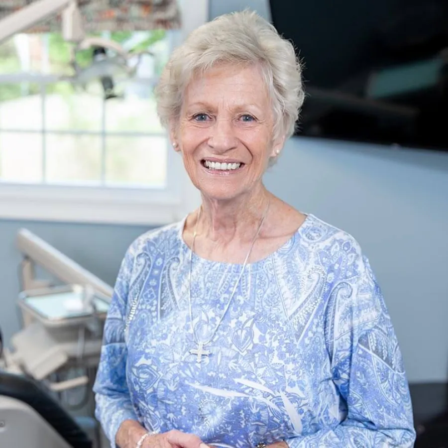 Smiling elderly woman with short white hair wearing a blue patterned top and cross necklace indoors