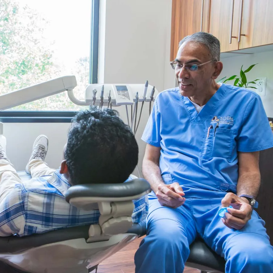 Dentist in blue scrubs talking to a boy in a dental chair with a girl waiting nearby in a bright clinic.