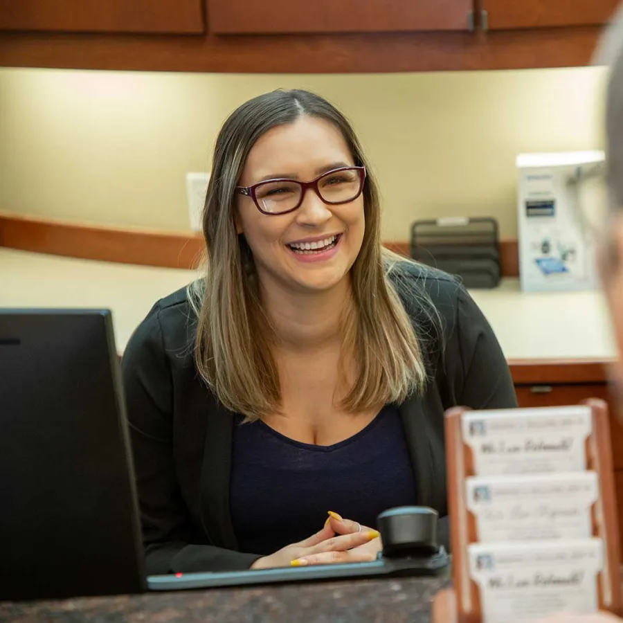 Smiling woman with glasses assists a customer at an office or service desk with computer and brochures.