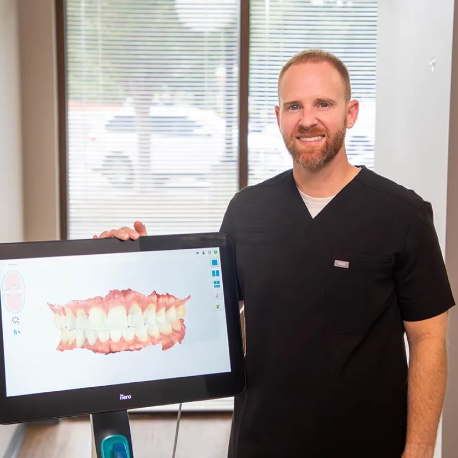 Male dentist in black scrubs displaying digital 3D dental scan on screen in bright office with window blinds.