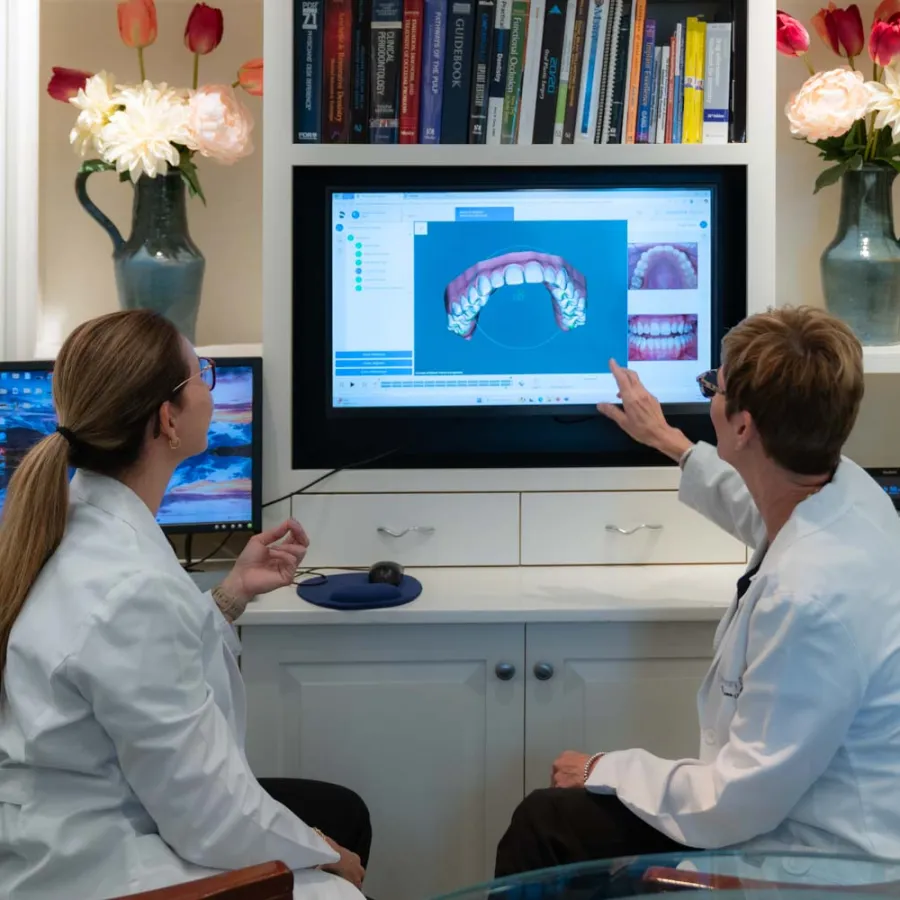 Two dentists in white coats discuss dental scan images on a computer screen in a bright office with flowers and books.