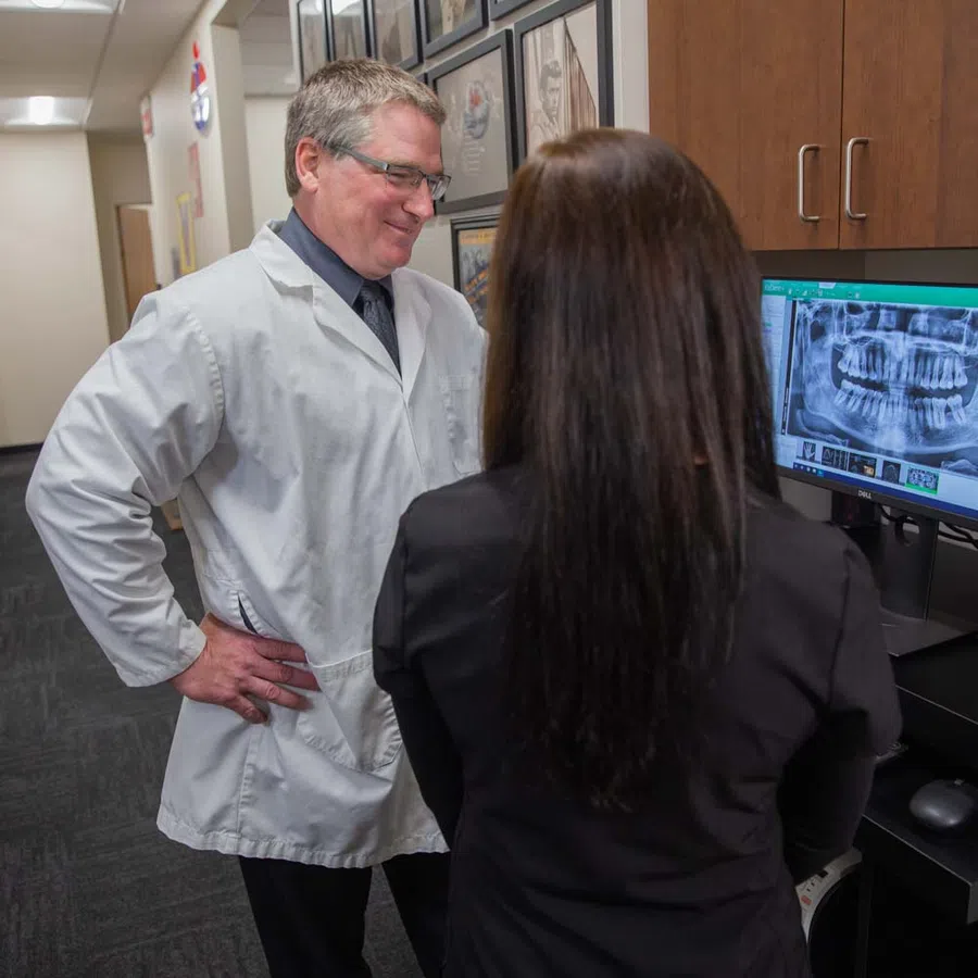 Dentist and patient reviewing a dental X-ray image on a computer screen in a clinic hallway.