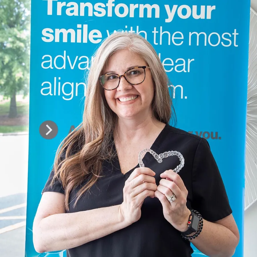 Smiling woman in glasses holding clear dental aligners shaped like a heart in front of a blue orthodontic sign.