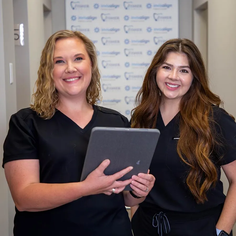 Two smiling female dental professionals in black scrubs holding a tablet in a dental clinic hallway.