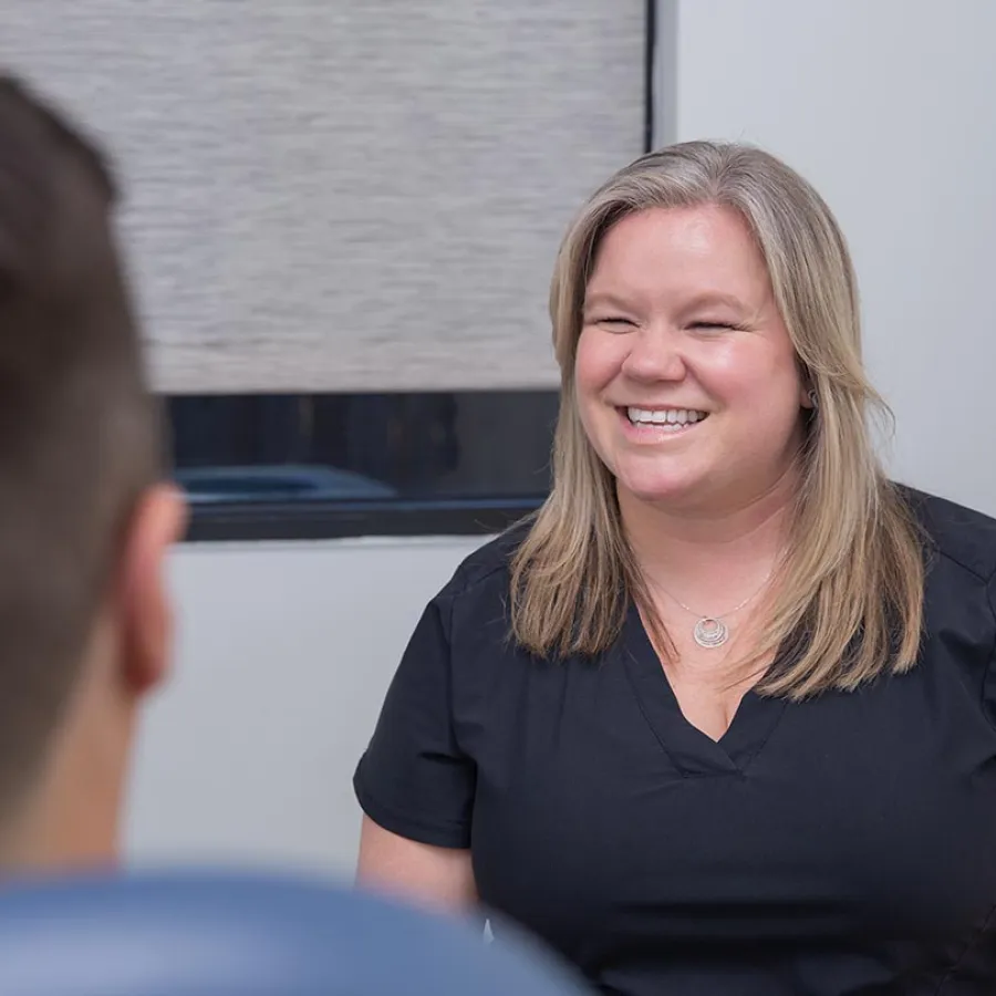 Smiling woman with blond hair in black shirt engaged in conversation indoors with blurred person.