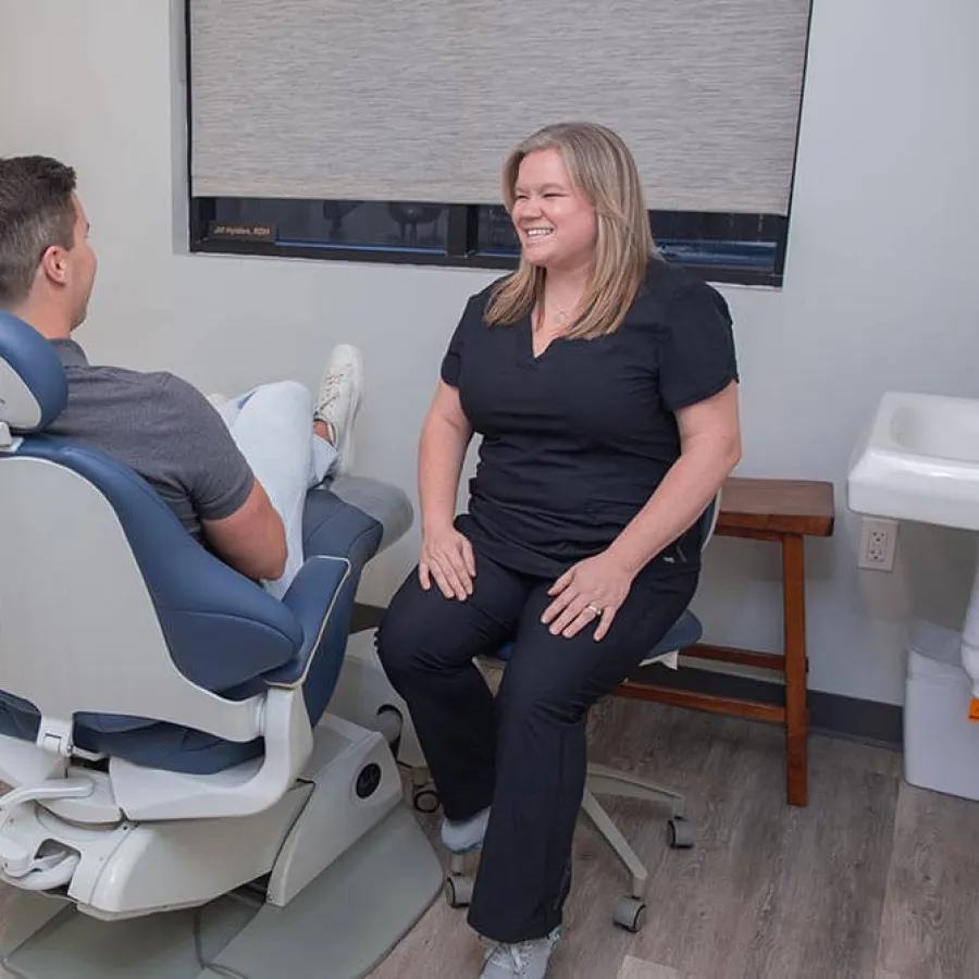 Dentist in black scrubs smiling and talking with patient in dental chair in a clinic room.