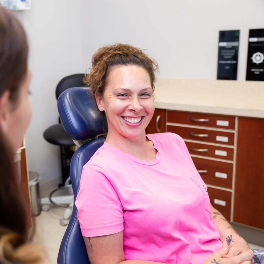 Smiling woman in a pink shirt sitting in a dental chair talking to a dentist in a clinic.