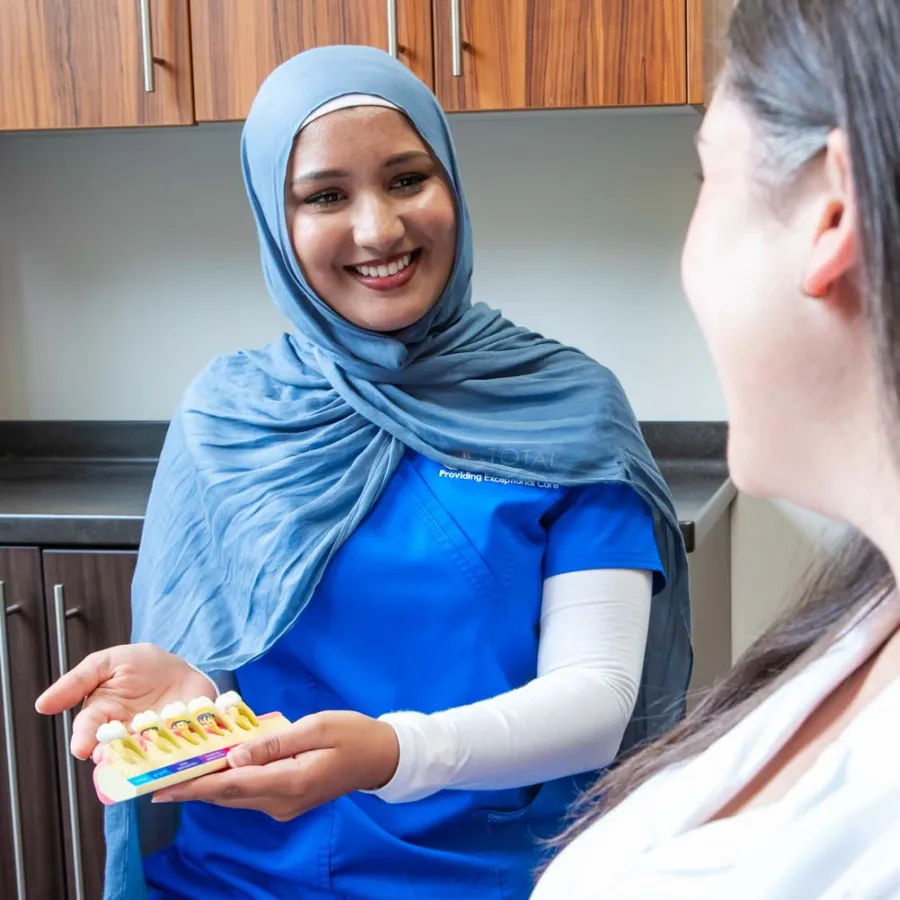Dental professional in blue scrubs and hijab explaining a teeth model to a patient in a clinic.