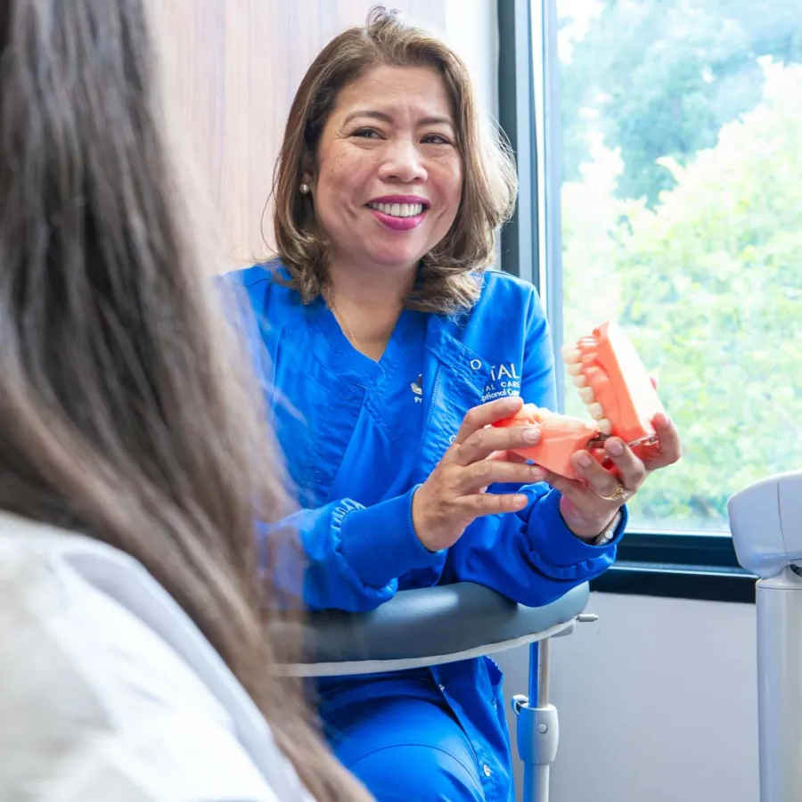 Dentist in blue scrubs smiling and showing a dental model to a patient in a consultation room.