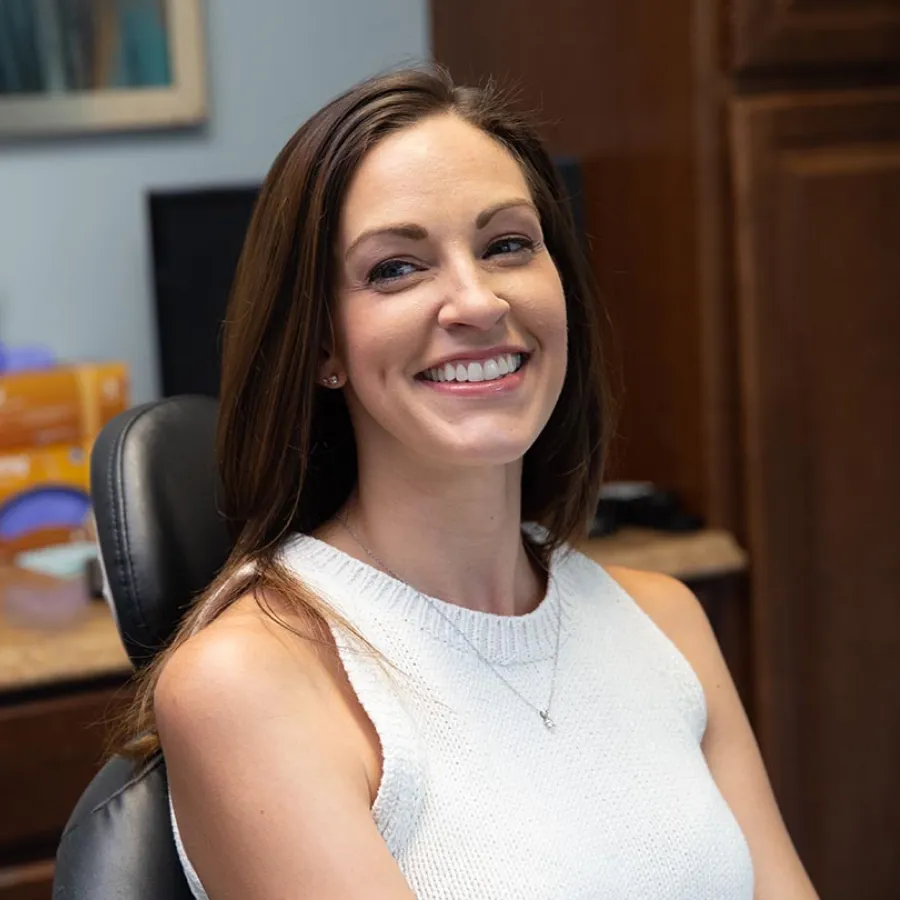 Smiling woman with long brown hair wearing a white sleeveless top sitting in an office chair indoors.