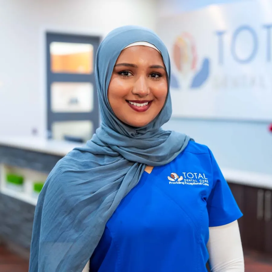 Smiling female dental professional in blue scrubs and hijab standing inside Total Dental Care clinic.