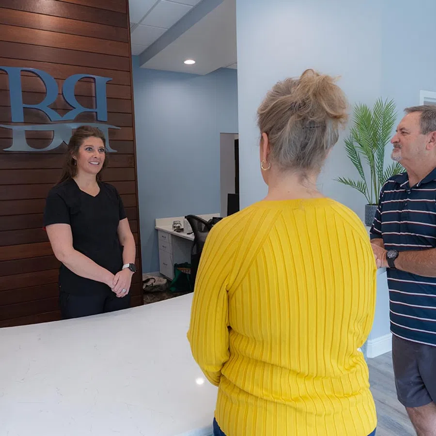 Receptionist in black talking to two visitors at a modern office front desk with wooden accent wall.