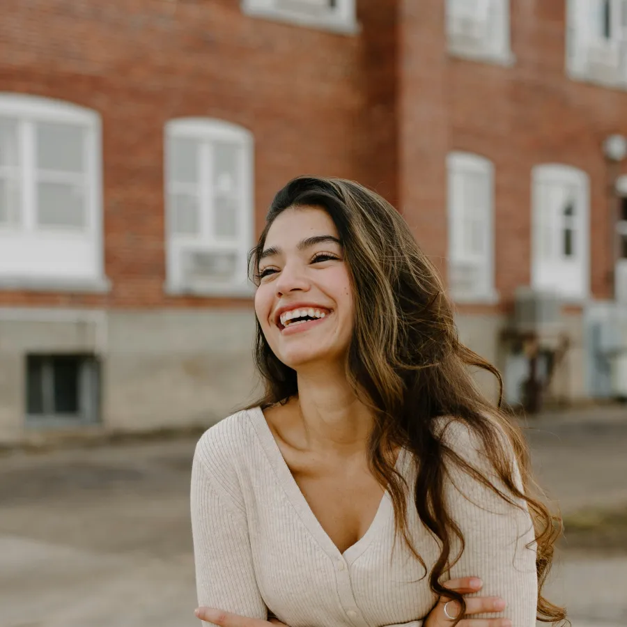 Smiling woman with long hair wearing a beige sweater and black pants standing outdoors by a brick building.