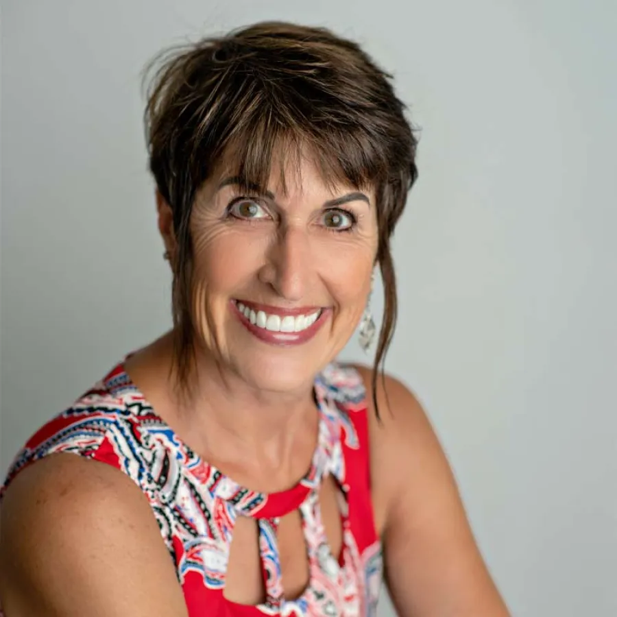 Smiling middle-aged woman with short brown hair wearing a red patterned sleeveless top against a neutral background
