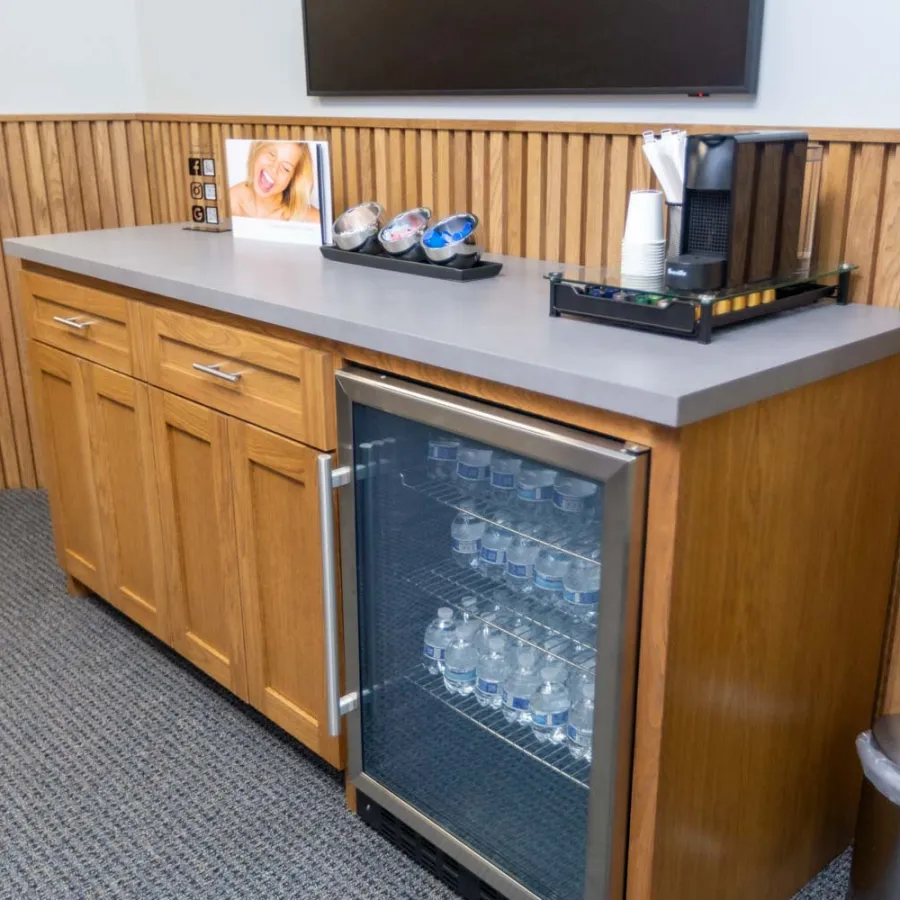 Wooden cabinet with mini fridge stocked with water bottles and coffee setup in a waiting area