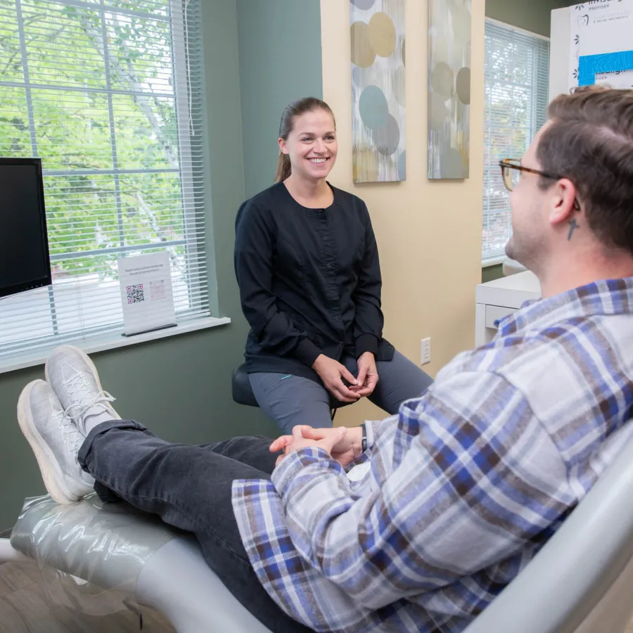Patient sitting in dental chair talking with smiling dental professional in a bright office with window and Invisalign signs.