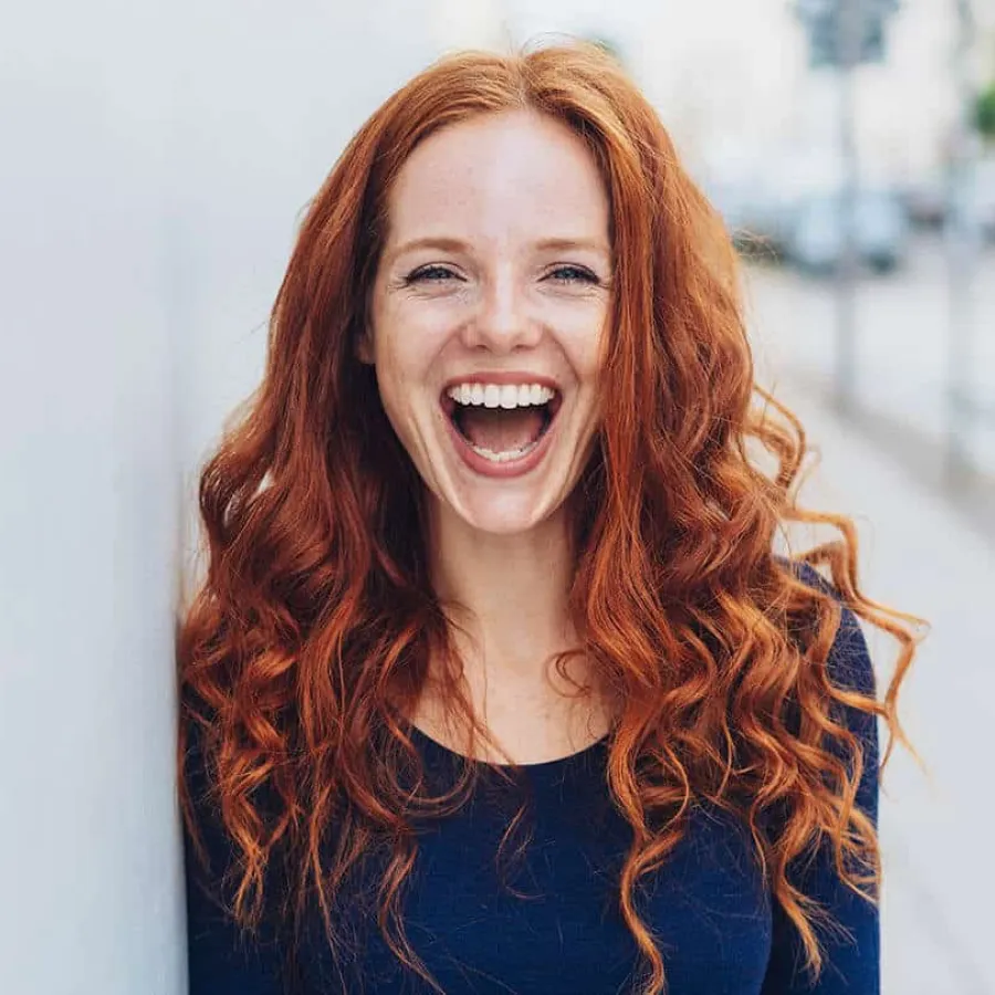 Young woman with long curly red hair smiling joyfully outdoors in casual dark blue top