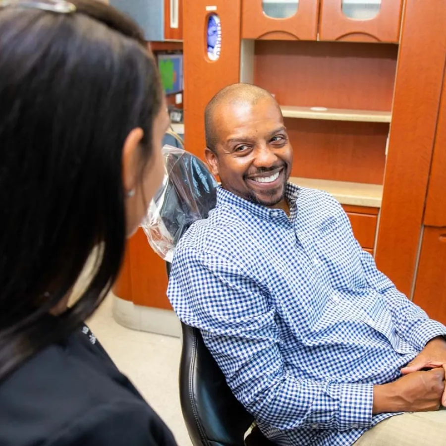 Man in checkered shirt smiling while sitting in a dental chair talking to a female dental professional.