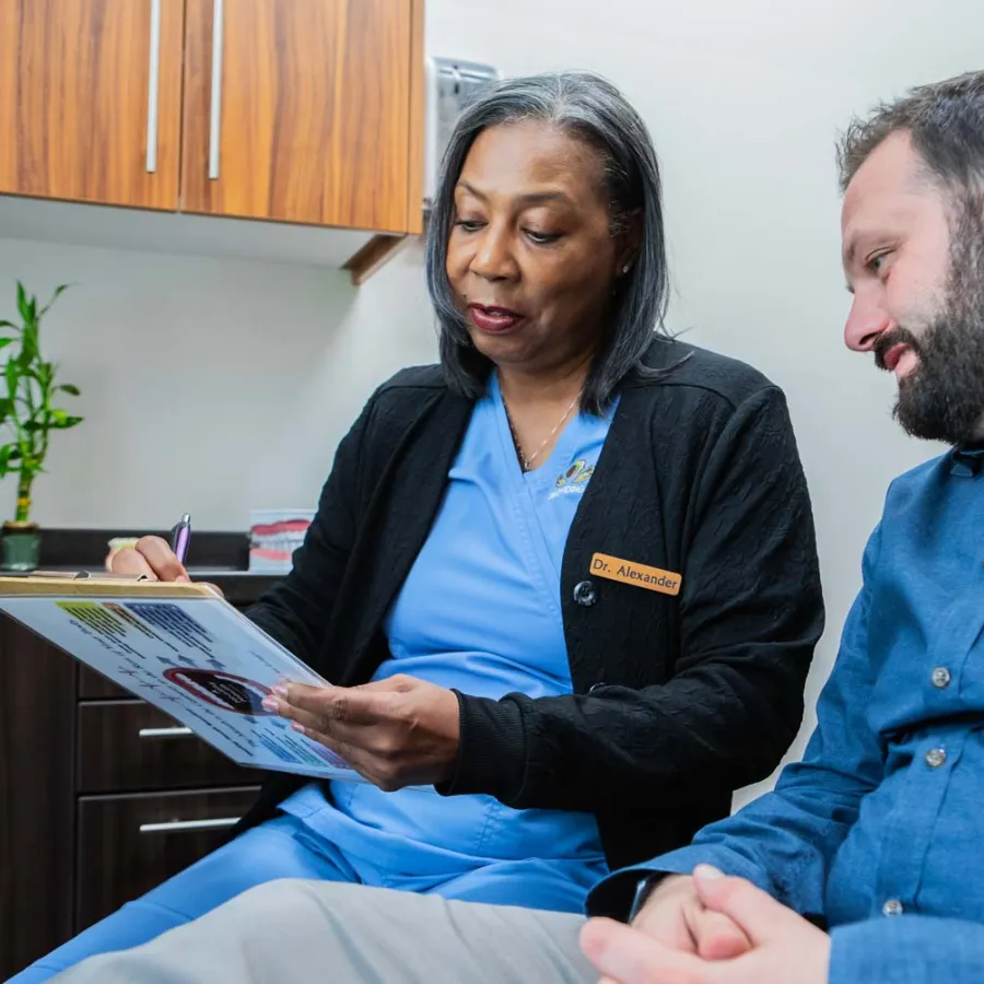 Female doctor in blue scrubs explaining medical information to male patient in clinic office.