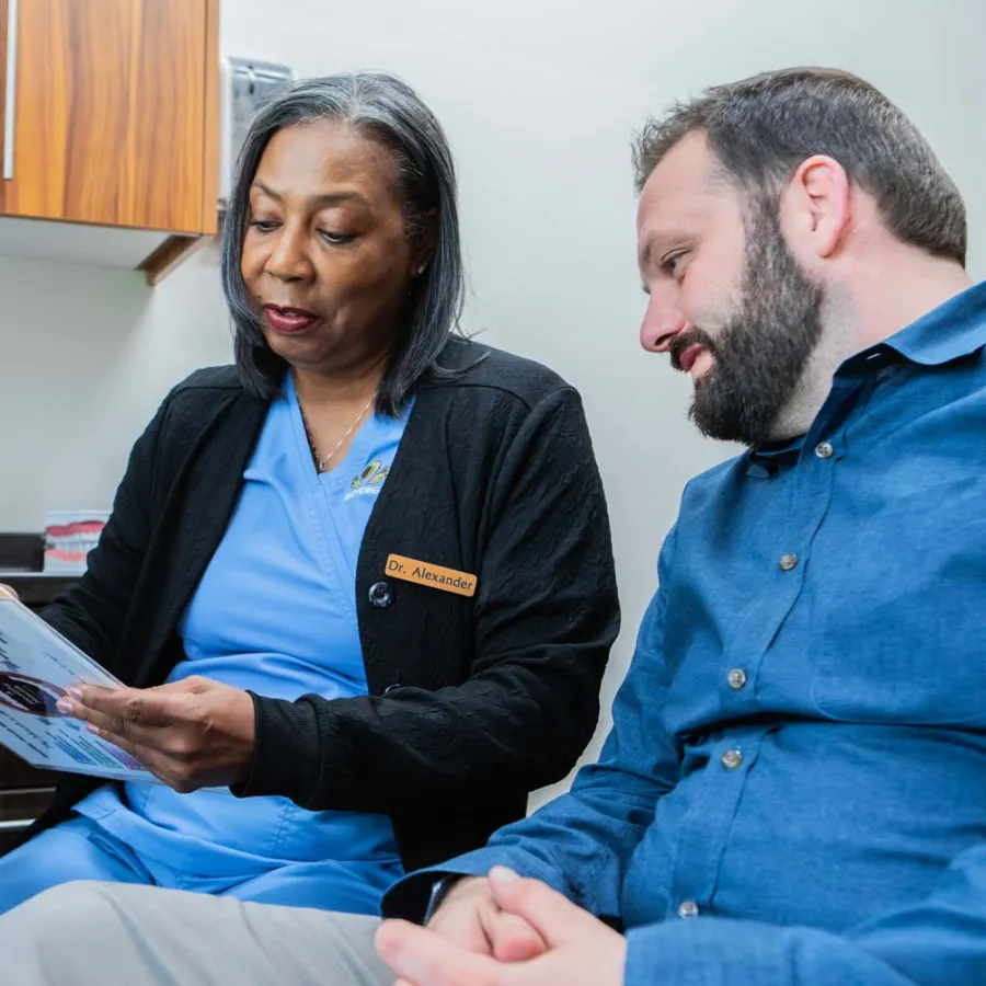Female doctor in blue scrubs explaining medical information to male patient in clinic office.