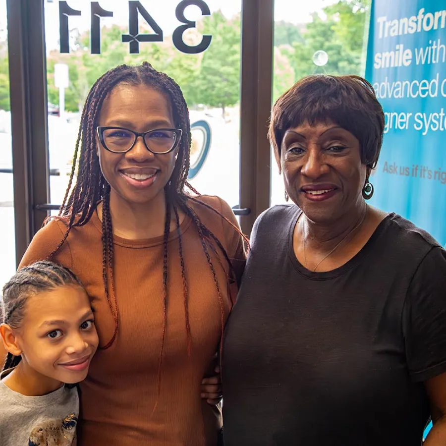 Three generations of smiling African American women standing indoors near a window with a sign in the background.