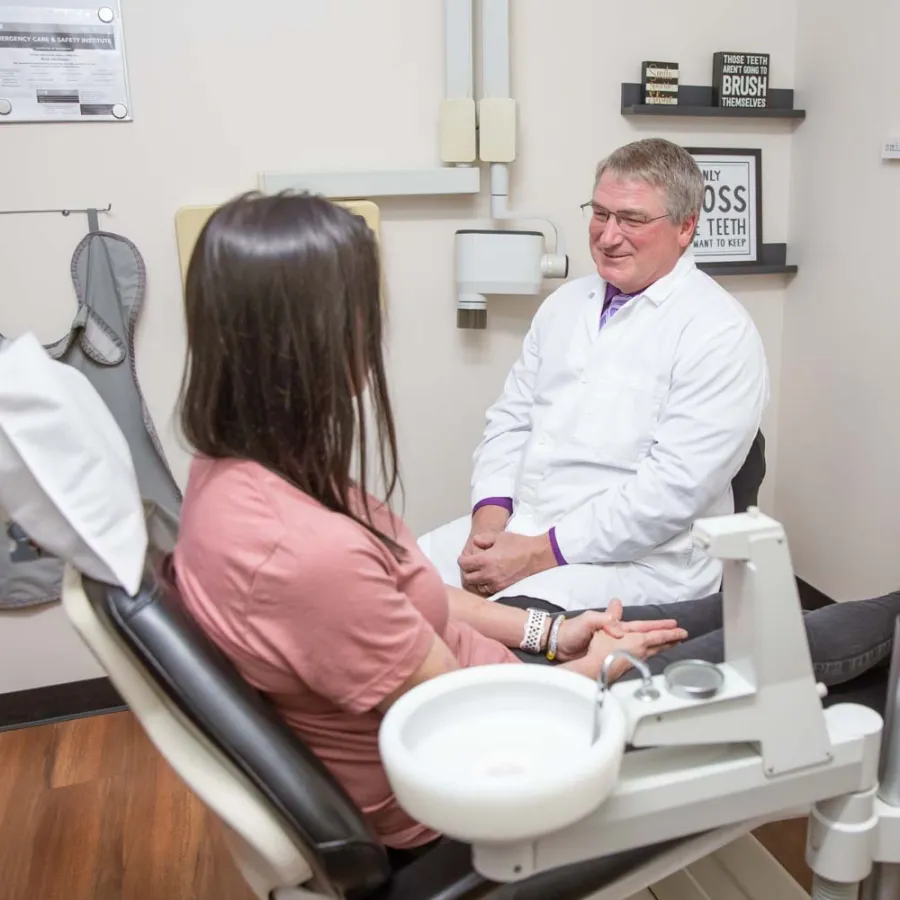 Dentist in white coat talking to a female patient sitting in a dental chair in a clinic room.