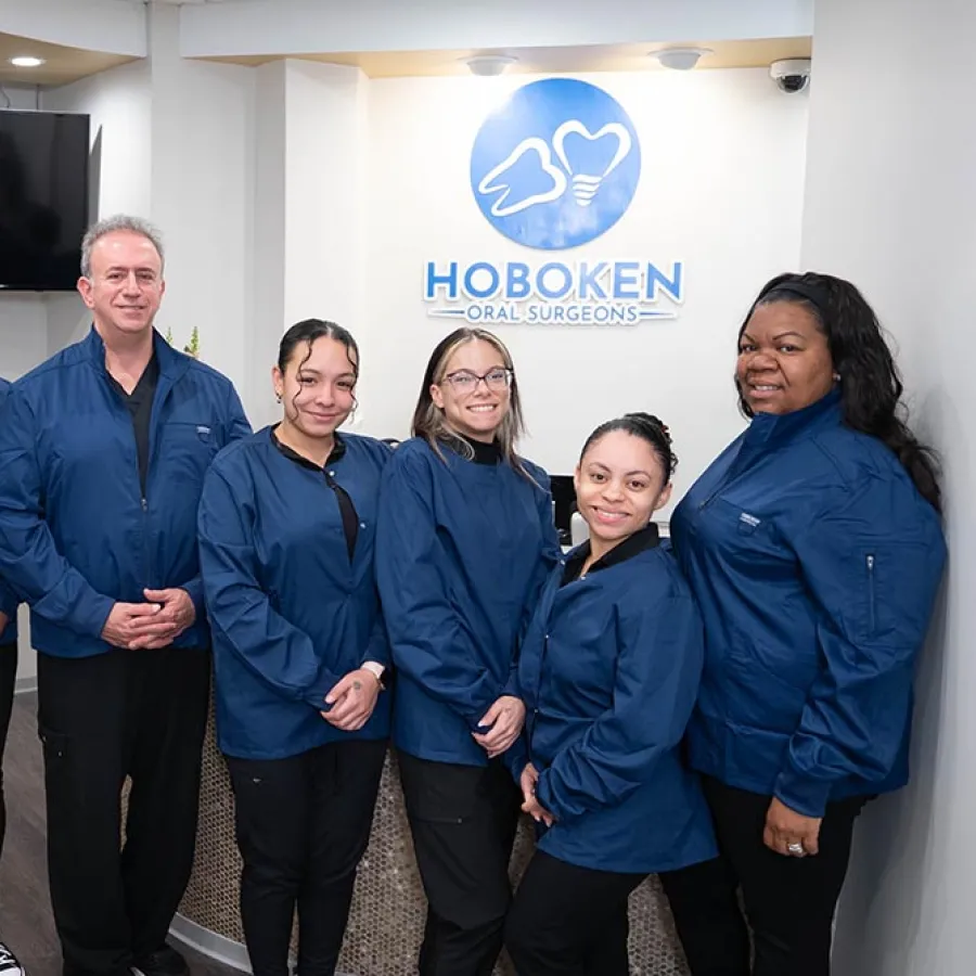 Group of seven Hoboken Oral Surgeons staff posing in blue uniforms in clinic lobby with logo behind them