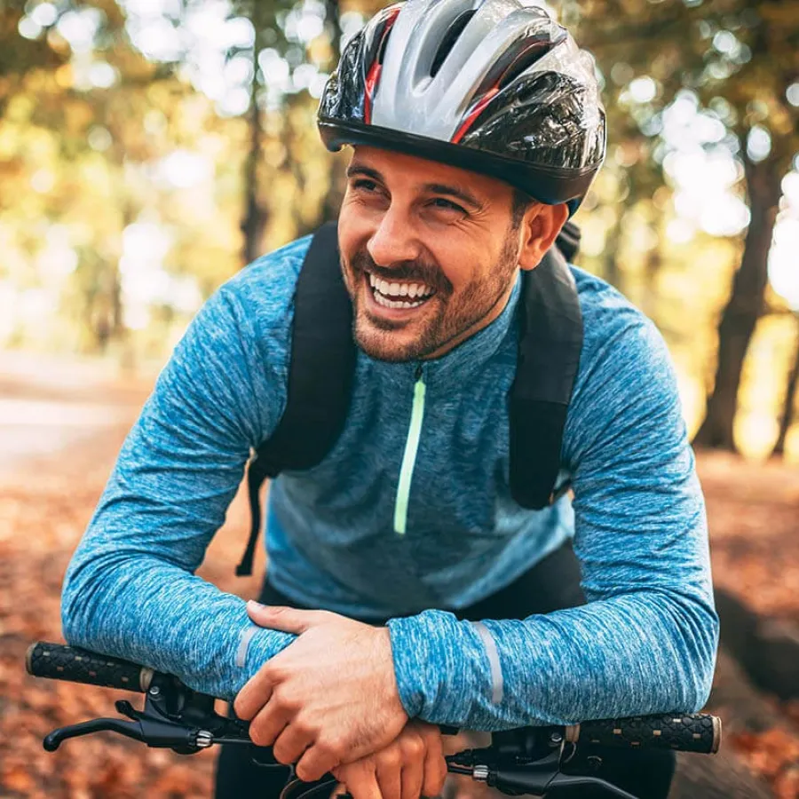 Smiling man in blue jacket and helmet leaning on bicycle handlebars in autumn forest.