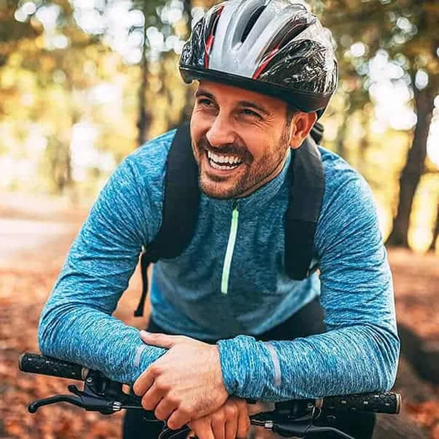 Smiling man in a blue jacket and helmet leaning on his bicycle handlebars in a forest with autumn leaves.