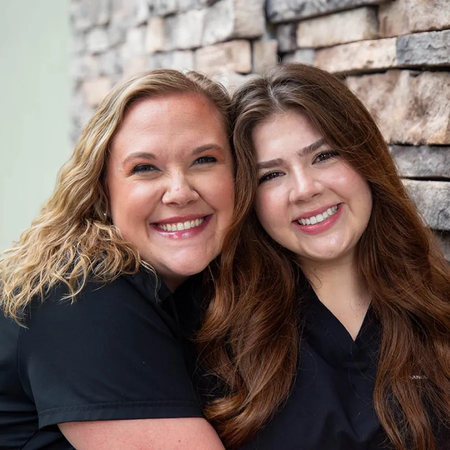 Two women wearing black scrubs smiling and embracing in front of a rustic stone wall background.