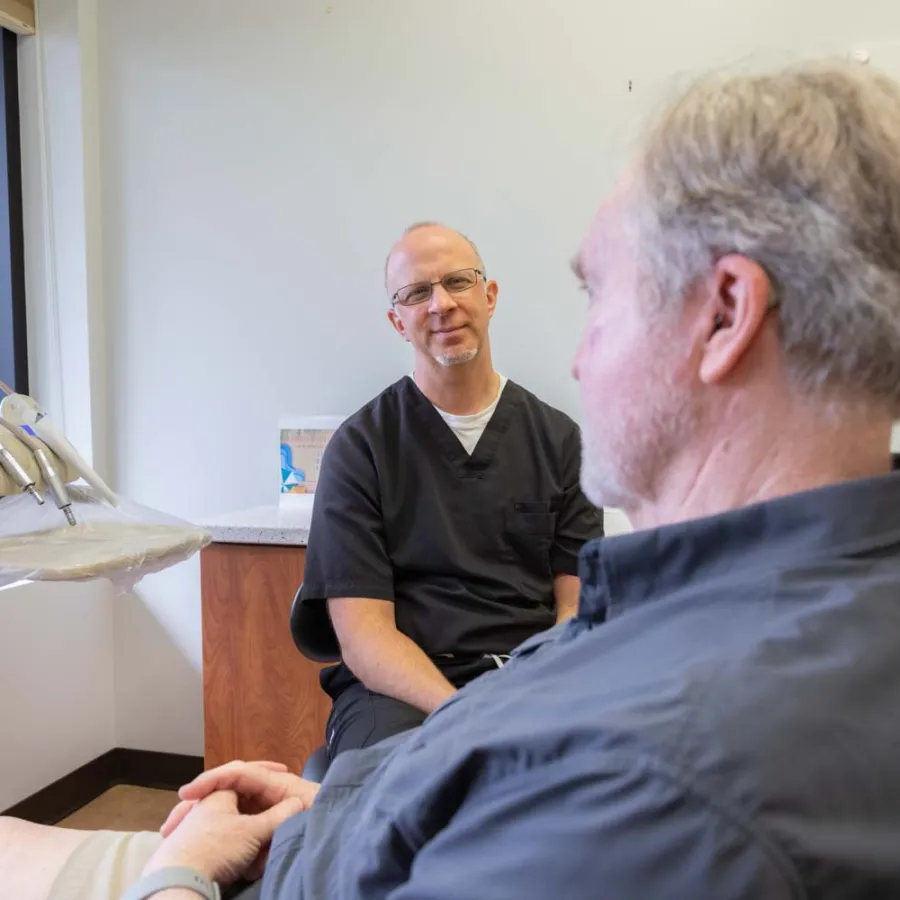 Dentist in black scrubs consulting with male patient in dental office with natural light