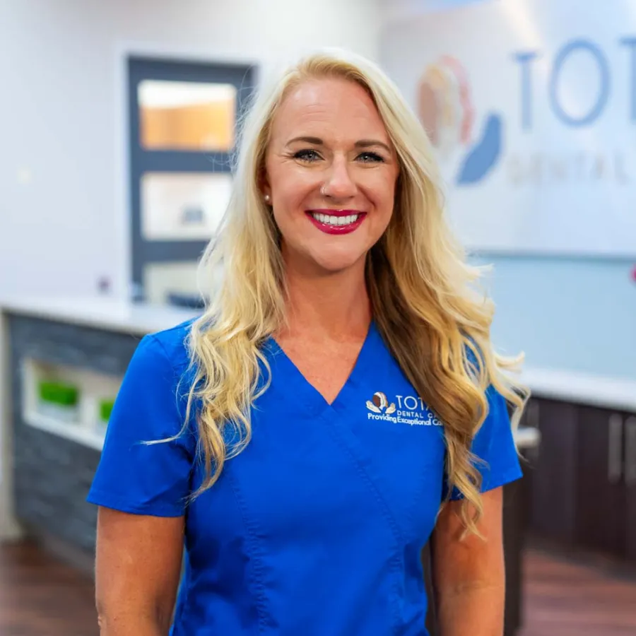 Smiling female dental professional in blue scrubs standing inside Total Dental Care clinic with logo in background.