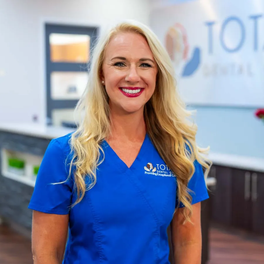 Smiling female dental professional in blue scrubs standing inside Total Dental Care clinic with logo in background.