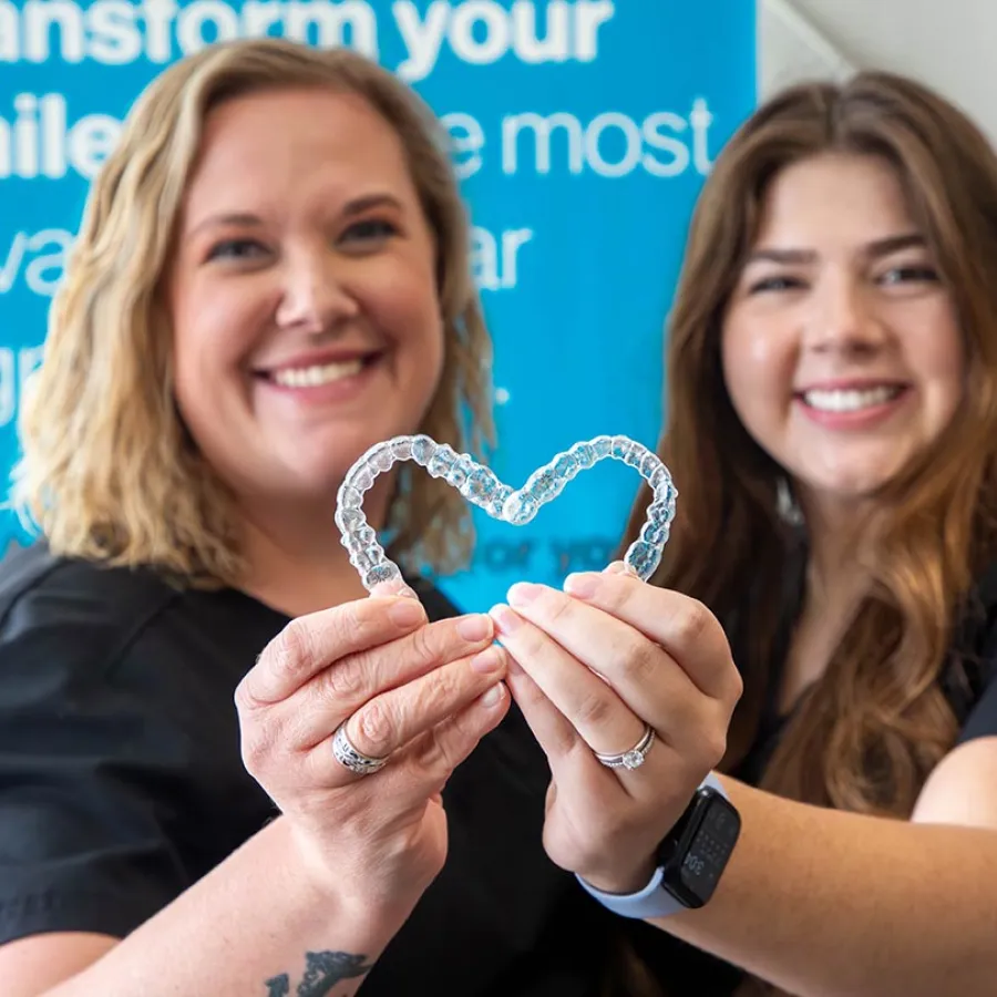Two smiling women holding a clear dental aligner shaped like a heart in a dental office.