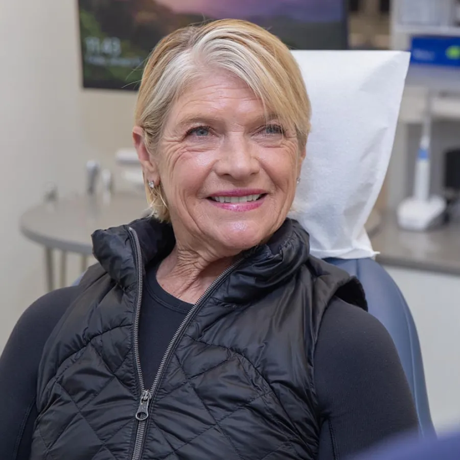 Smiling elderly woman with short blonde hair wearing a black vest sitting in a medical or dental office chair.
