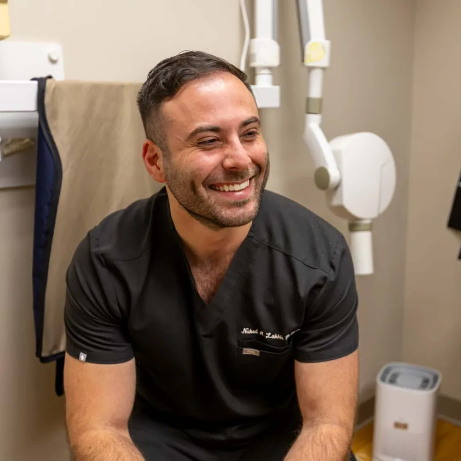 Smiling male dentist in black scrubs sitting in a modern dental office with equipment and sink in background