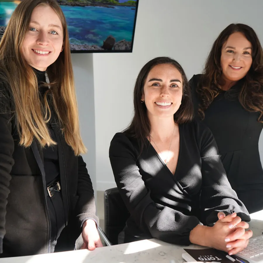 Three professional women smiling in an office setting with a ocean landscape screen in the background