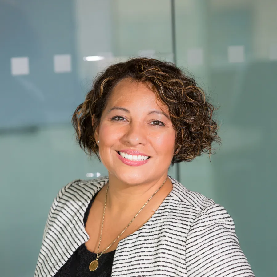 Smiling confident woman with curly hair wearing a black top and striped blazer in an office setting.