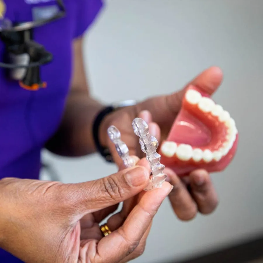 Smiling young woman inserting clear dental aligners over her teeth indoors with warm lights in the background