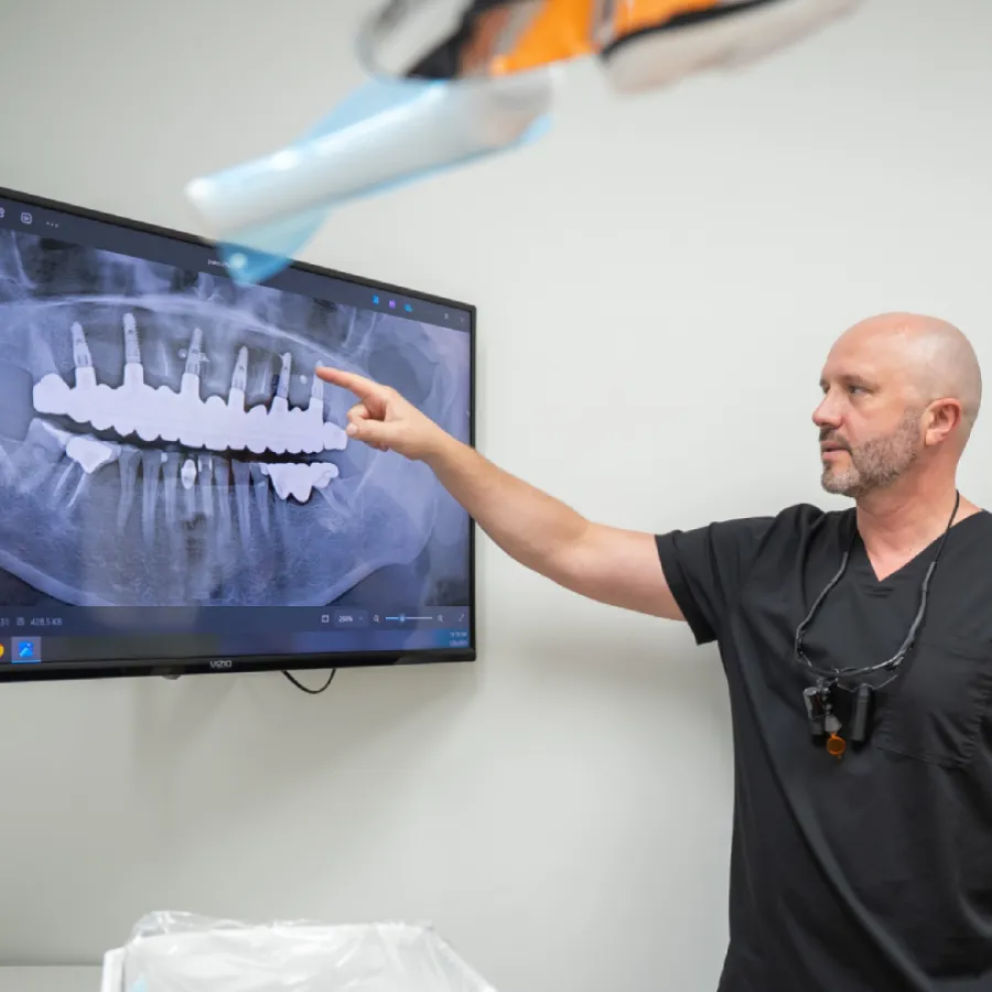 Dentist pointing at dental X-ray displayed on monitor showing teeth and implants in a clinical setting.