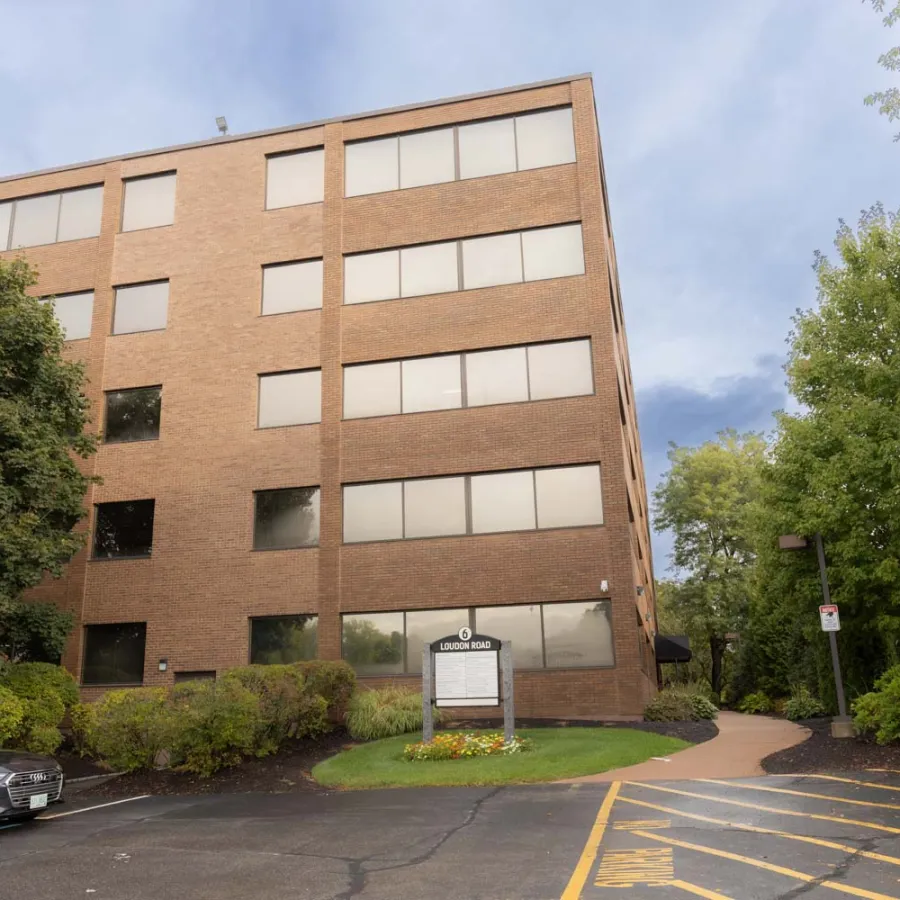 Four-story brick office building with parked cars, landscaping, and a clear sky background.