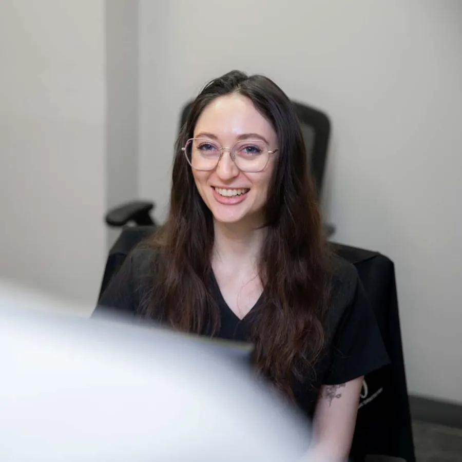 Smiling woman with long brown hair and glasses sitting in an office chair in a modern workspace.
