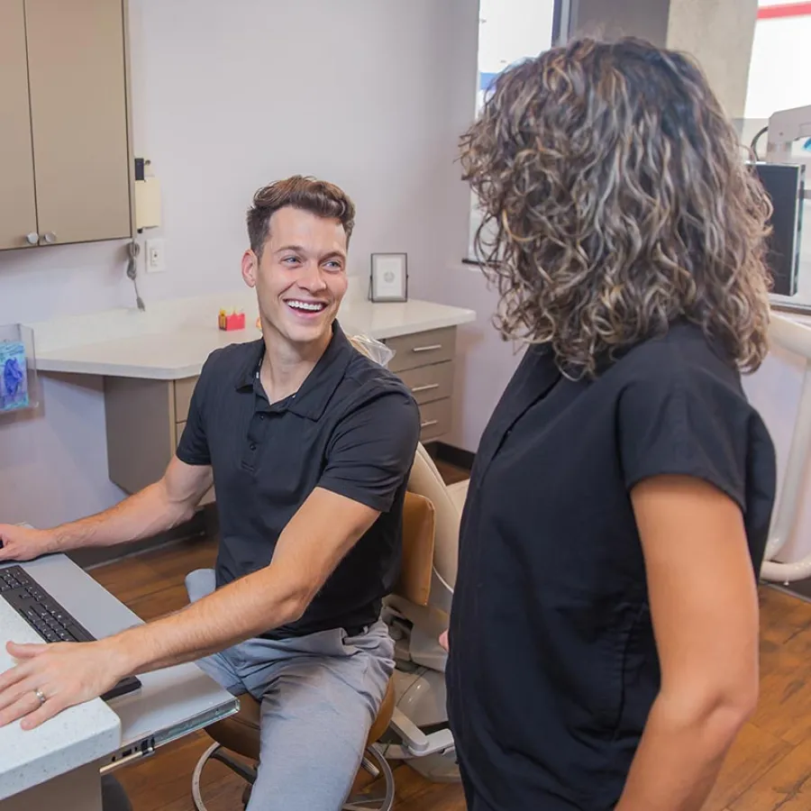 Smiling man sitting at a desk with a computer talks to a standing woman in a casual office setting.