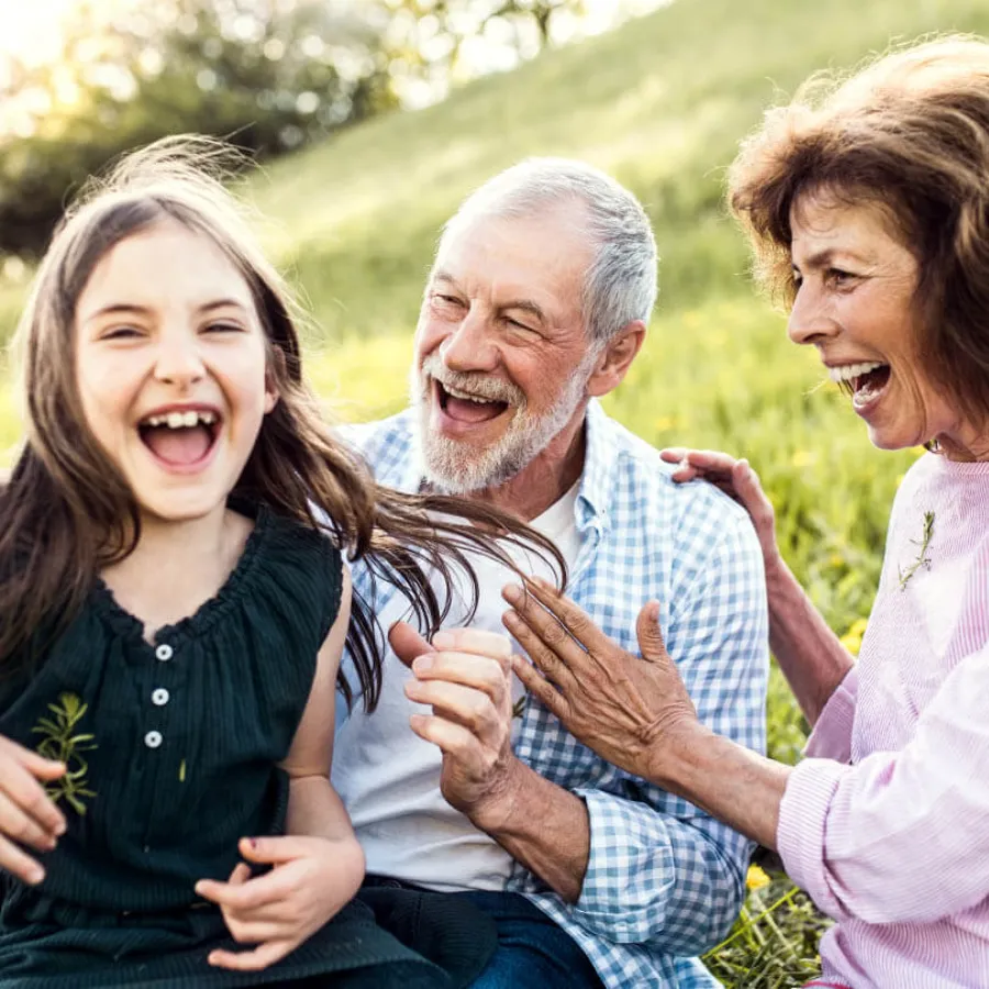 Happy grandparents laughing and playing outdoors with their granddaughter in a sunny green meadow.