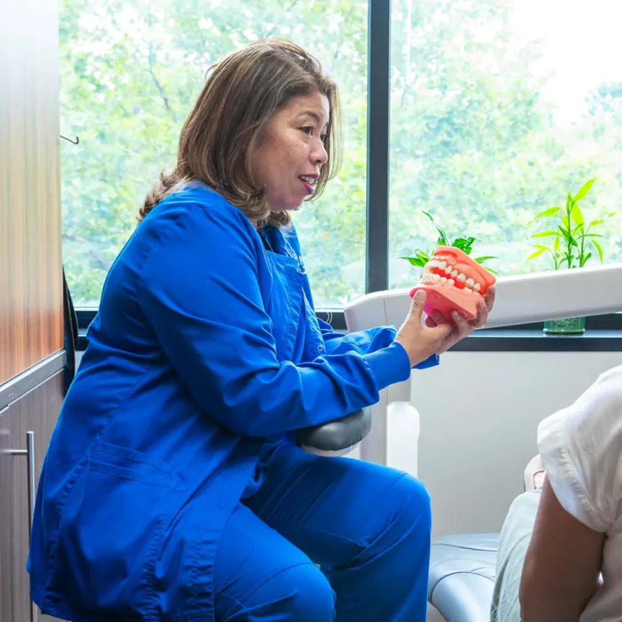 Dentist in blue scrubs explaining dental care using a teeth model to patient in dental chair near a window.