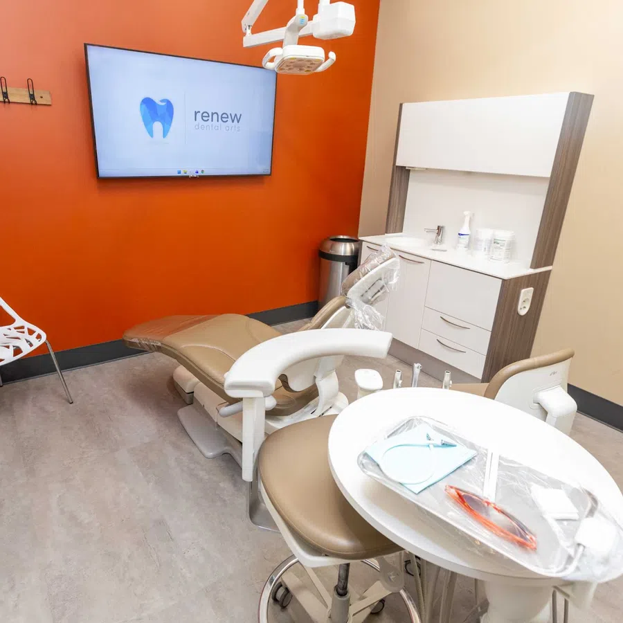 Modern dental office with beige chair, orange accent wall, white shelving, and screen displaying Renew Dental Arts logo.